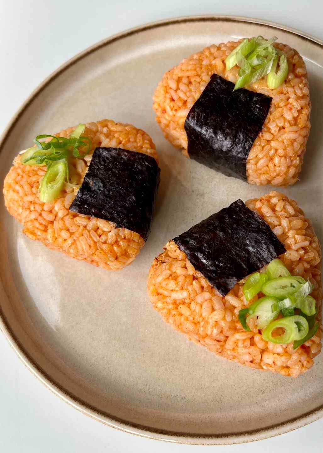 Three triangular gochujang onigiri Korean rice balls coated in a vibrant red-orange gochujang seasoning, each wrapped in a strip of nori seaweed, sliced open to show the tuna mayonnaise filling, garnished with finely sliced spring onions on a white plate.
