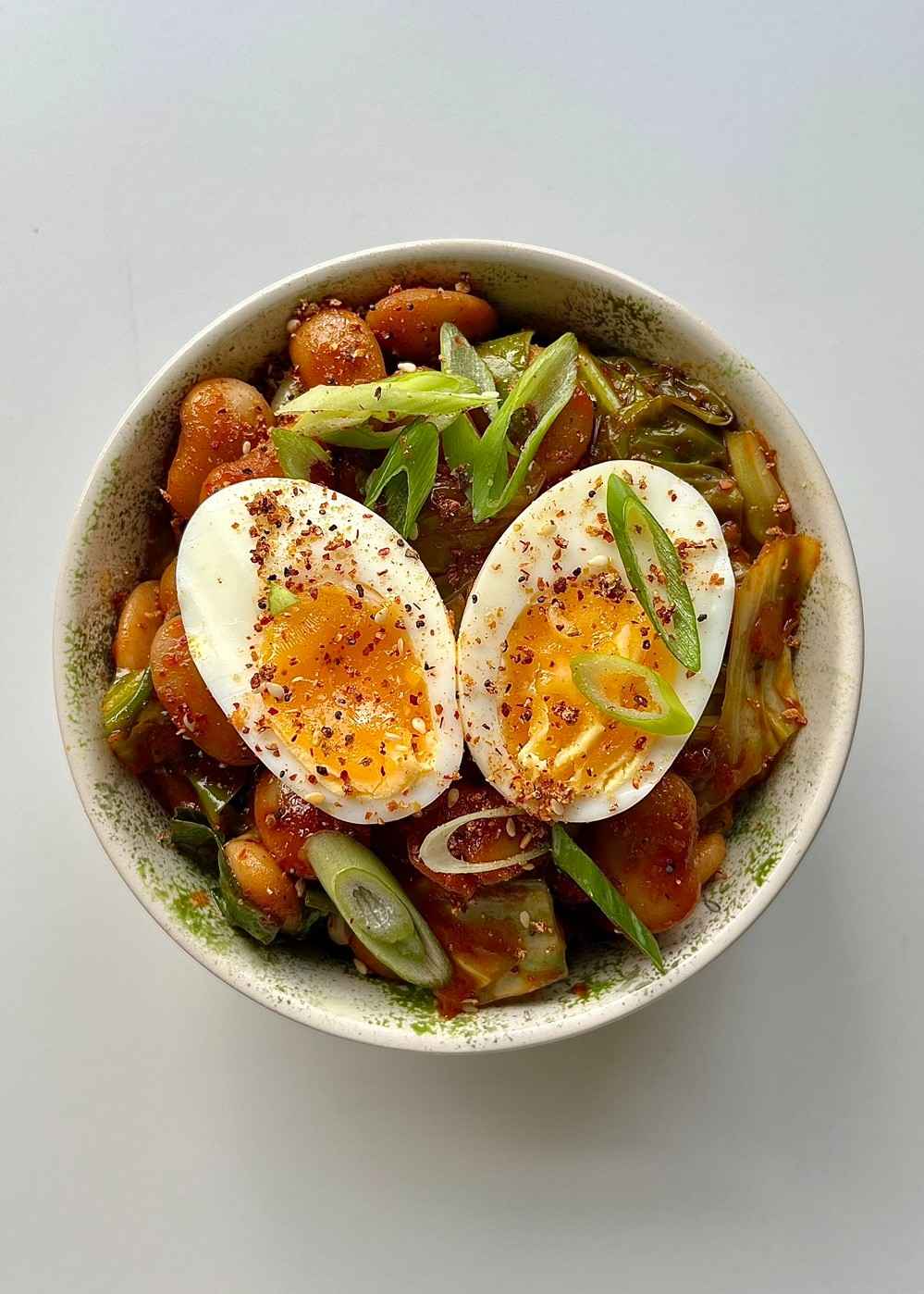 A bowl of Korean-style butter beans in a glossy red gochujang sauce on a bed of white rice, topped with shredded cabbage, sliced spring onions, and a soft-boiled egg cut in half showing the jammy yolk.
