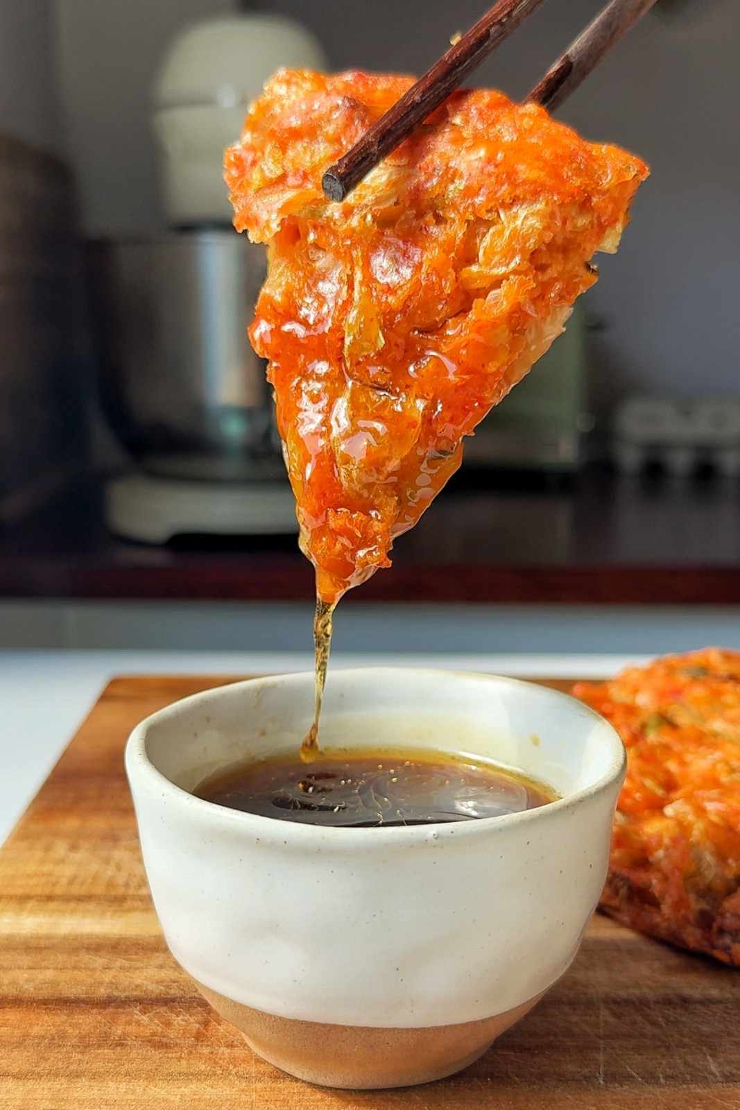 A slice of golden-brown kimchijeon traybake being dipped into a small bowl of soy-vinegar dipping sauce, with crispy edges and visible pieces of red kimchi and green scallions throughout the savory pancake. Alt text.