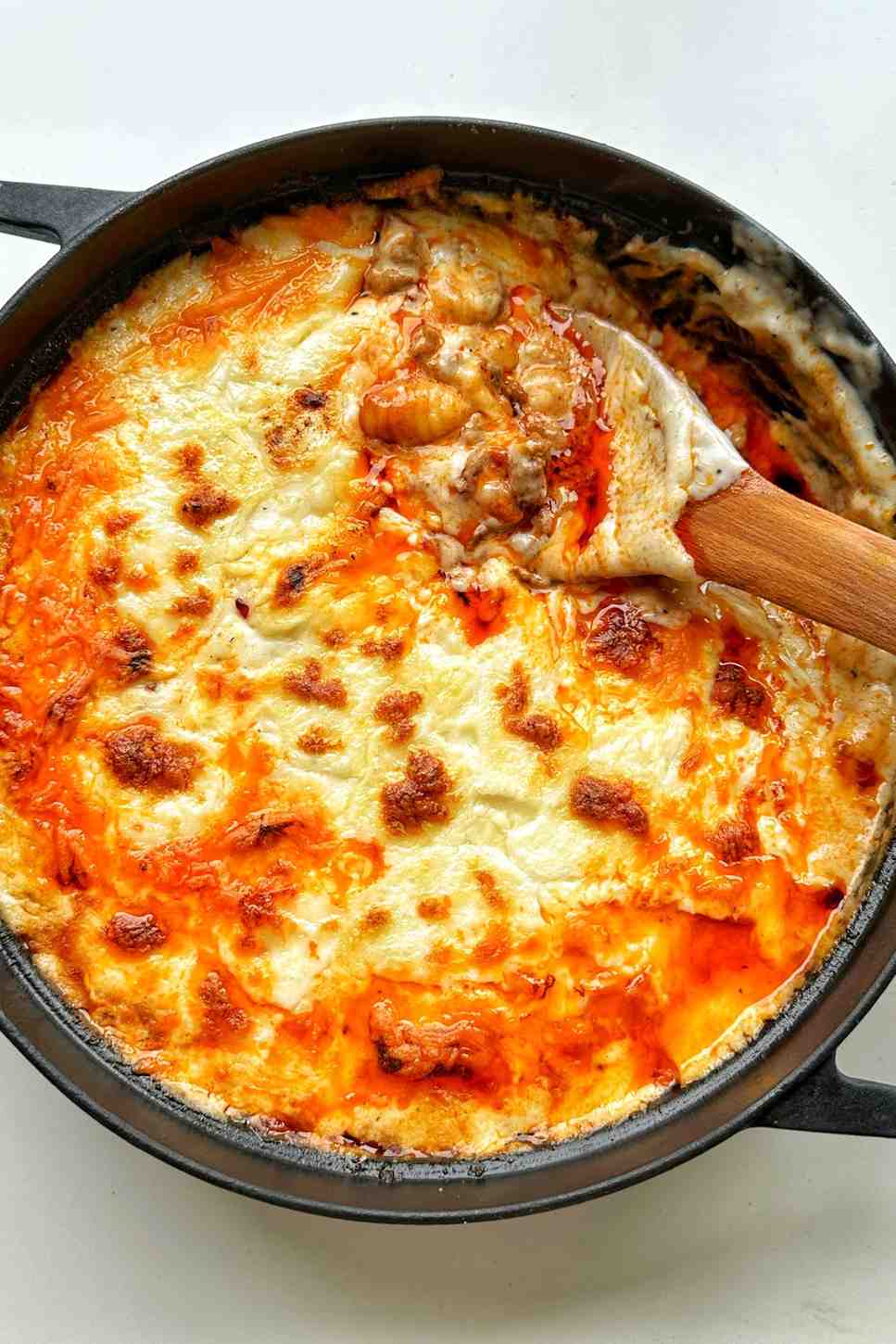 A black casserole dish filled with gnocchi lasagne with gochujang showing layers of beef ragu, soft pillowy gnocchi, creamy béchamel sauce, and a golden bubbling top of melted mozzarella and torn fresh mozzarella, with a wooden spoon scooping out a portion to reveal the saucy interior.