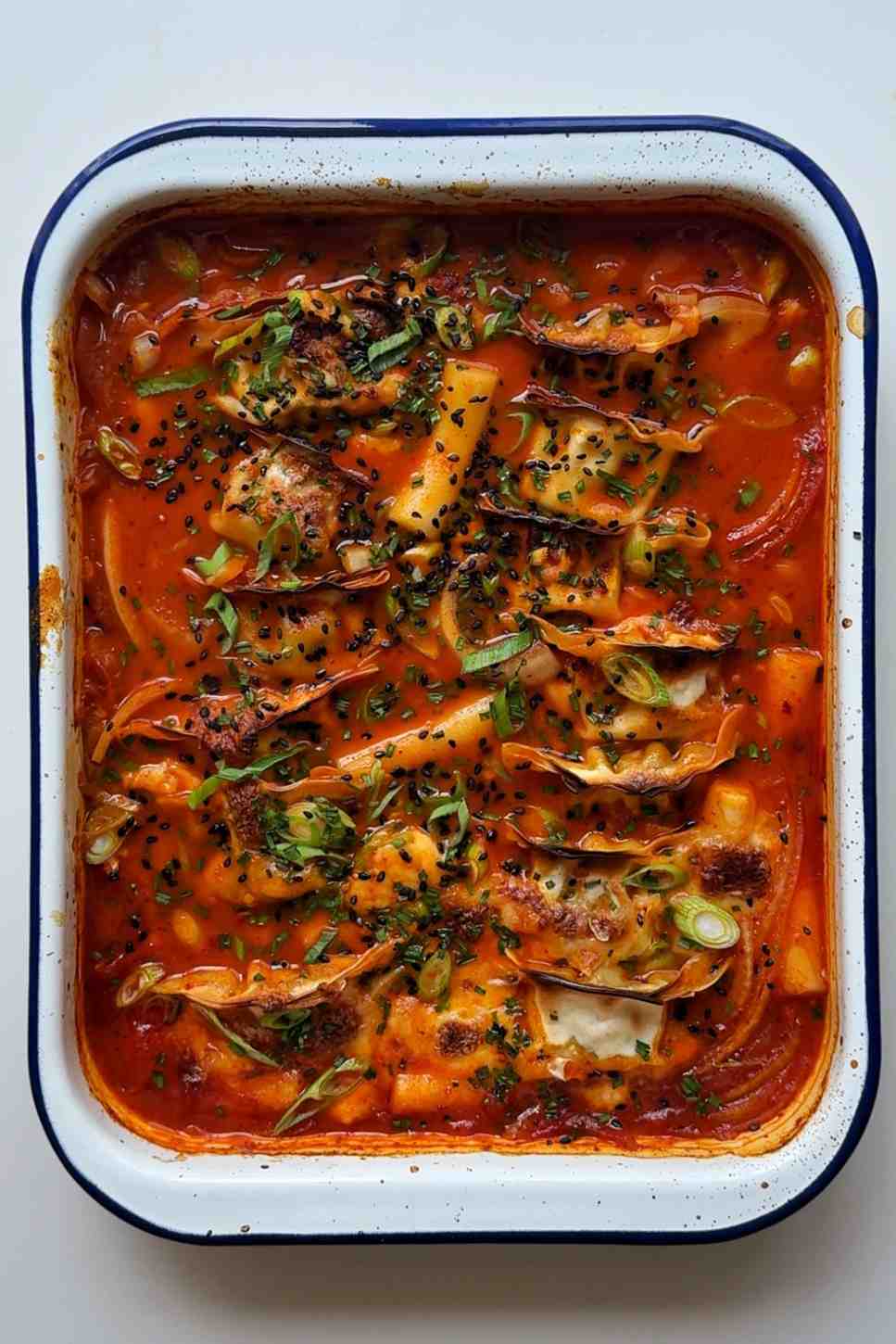 A white baking tray of dumpling tteokbokki bake with dumplings nestled into a glossy red gochujang sauce surrounded by chewy white rice cakes, topped with melted golden mozzarella, and garnished with sliced spring onions, chives, and black sesame seeds.