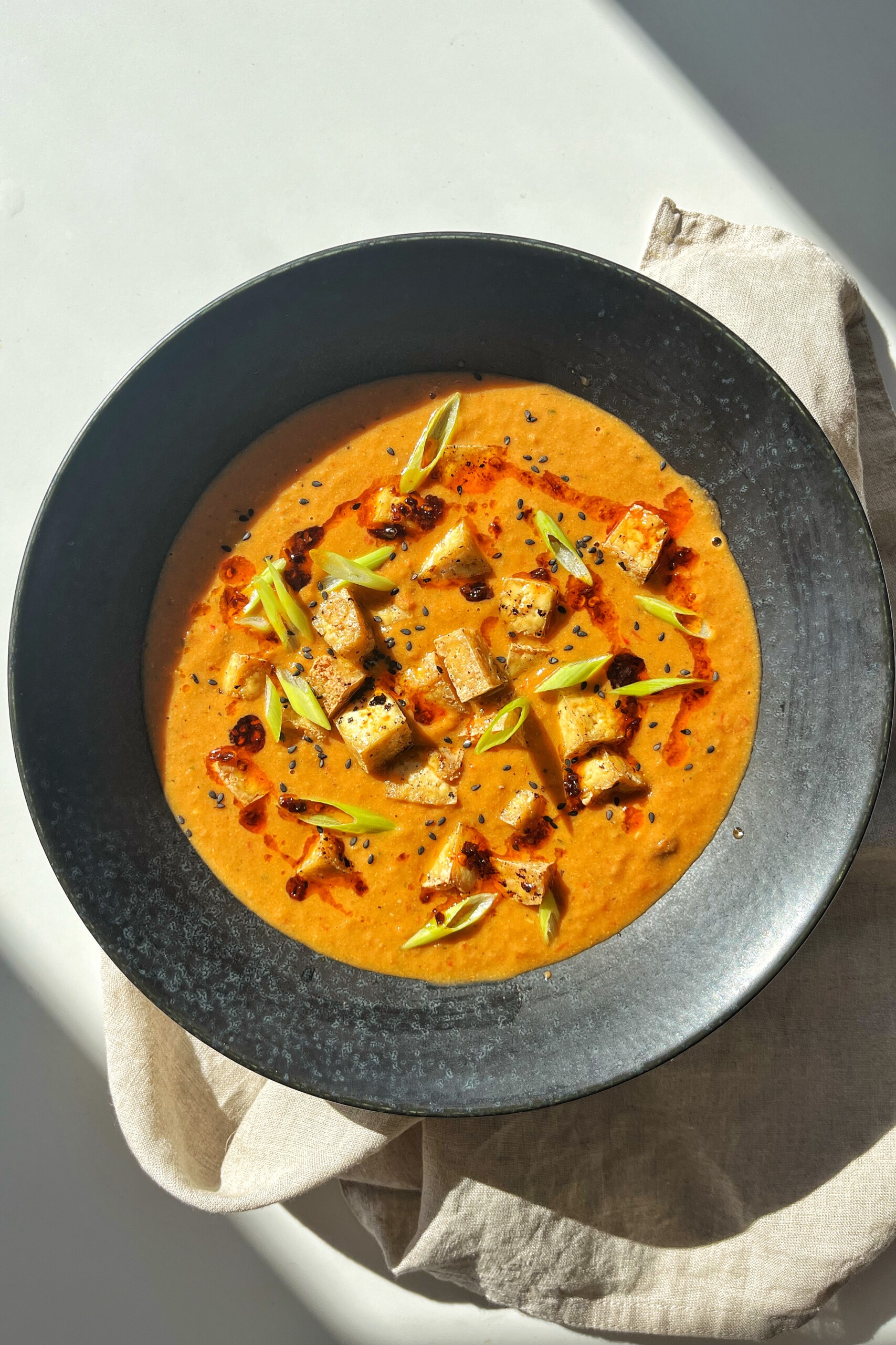Overhead bowl of creamy tofu tomato soup topped with crispy tofu cubes, sliced scallions, chili oil, and black sesame seeds.