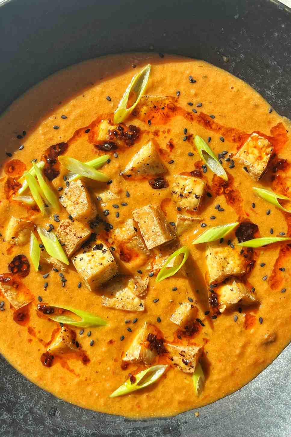 Close-up of tofu tomato soup in a dark bowl with crispy air-fried tofu cubes, chili oil, sliced scallions, and black sesame seeds in a creamy orange-red tomato base.