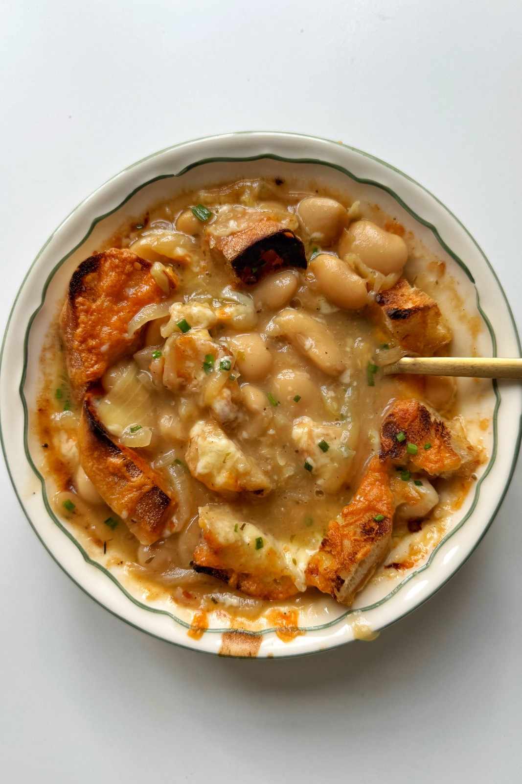 Overhead bowl of French onion style butter bean soup with plump white beans in a caramelized onion broth, topped with charred cheesy baguette slices and fresh chives.