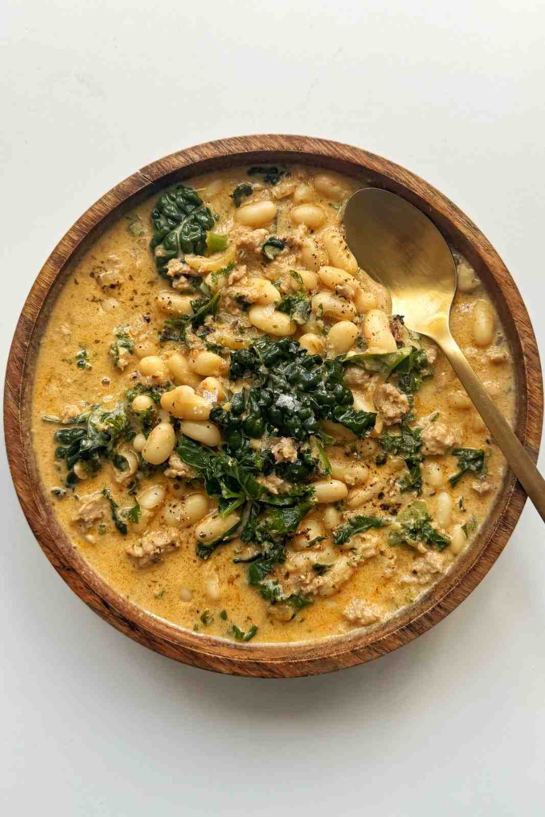 Overhead wooden bowl of kale and white bean sausage soup with Italian sausage, plump white beans, and wilted kale in a creamy golden broth, with a gold spoon.