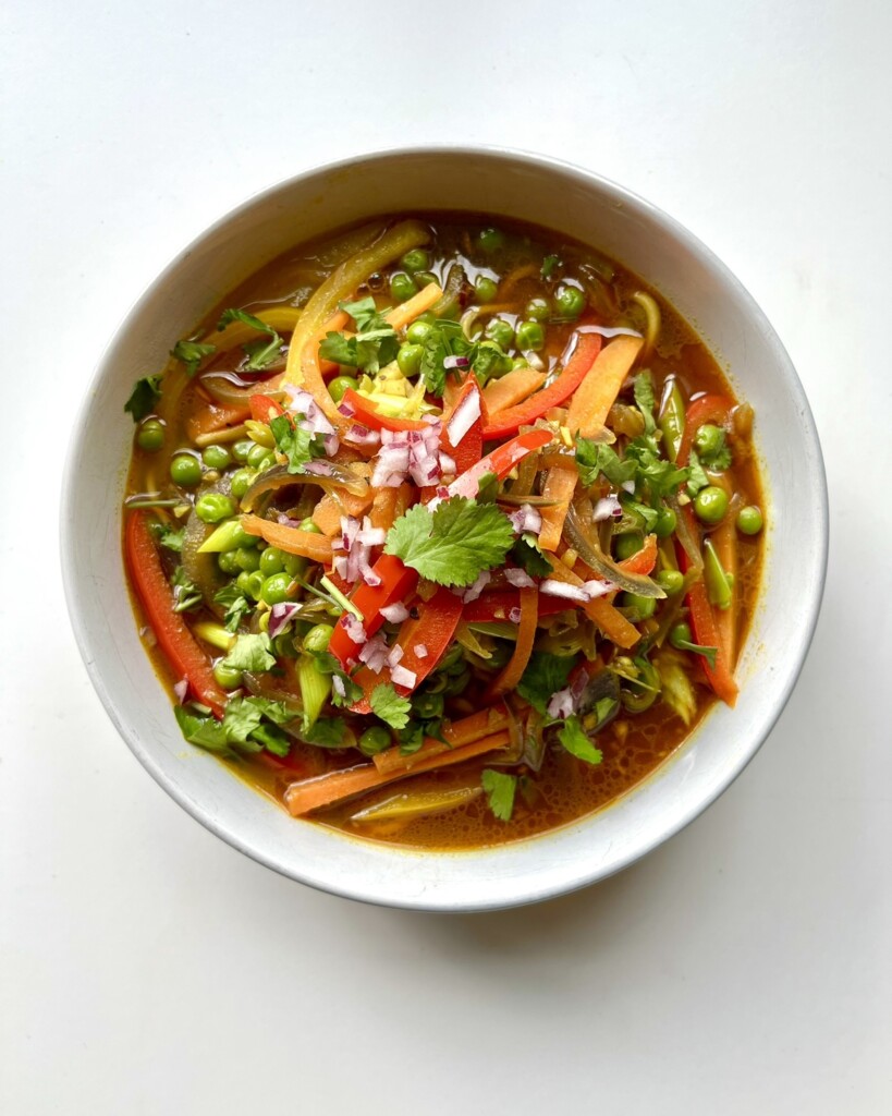 Overhead bowl of vegetable thukpa, Tibetan noodle soup, with egg noodles, peas, carrots, and peppers in a spiced broth, topped with scallions and cilantro.