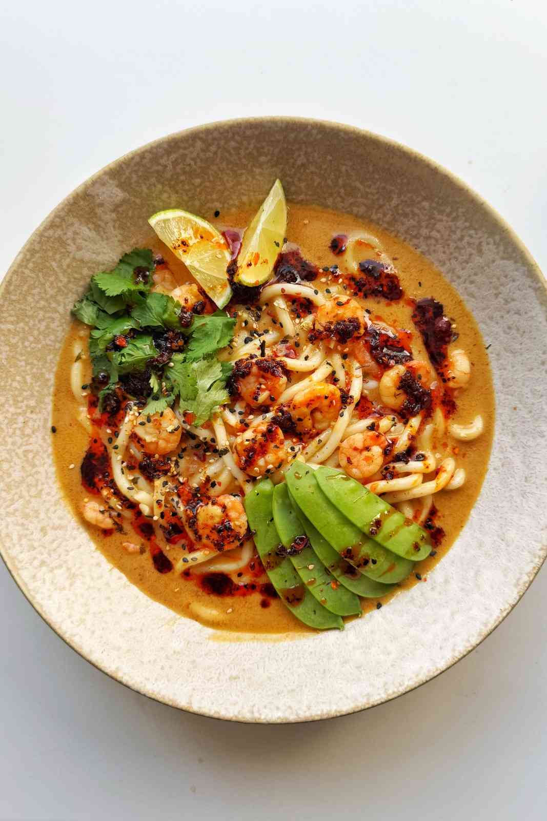 Overhead bowl of shrimp ramen noodle soup in a creamy coconut and curry broth, topped with shrimp, snow peas, cilantro, lime wedges, and chili oil.