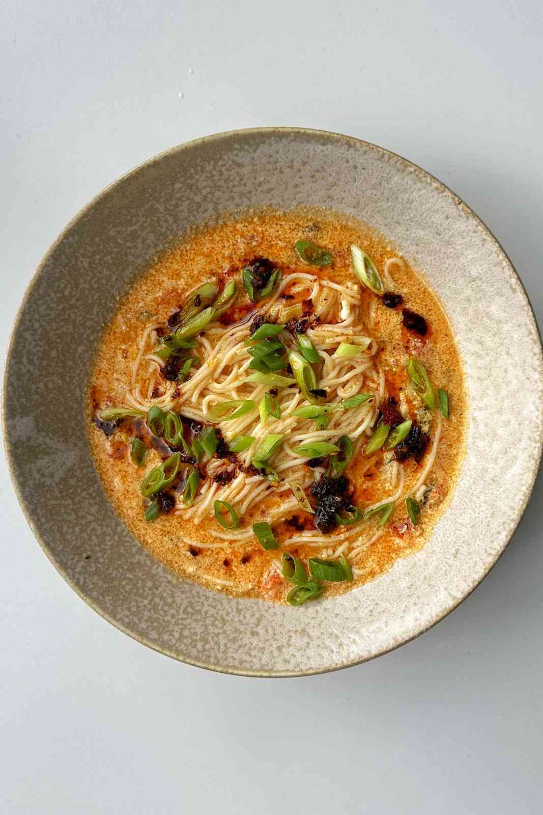 Overhead bowl of tomato egg drop noodle soup with thin noodles in a spicy orange-red tomato broth, topped with sliced scallions and chili oil.