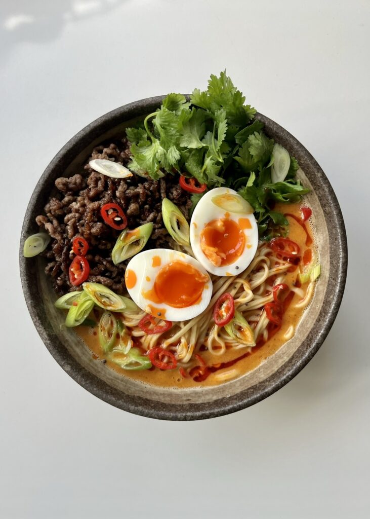 Overhead bowl of 15-minute spicy beef noodle soup with crispy ground beef, jammy eggs, noodles in a peanut broth, topped with cilantro, sliced scallions, and red chili.
