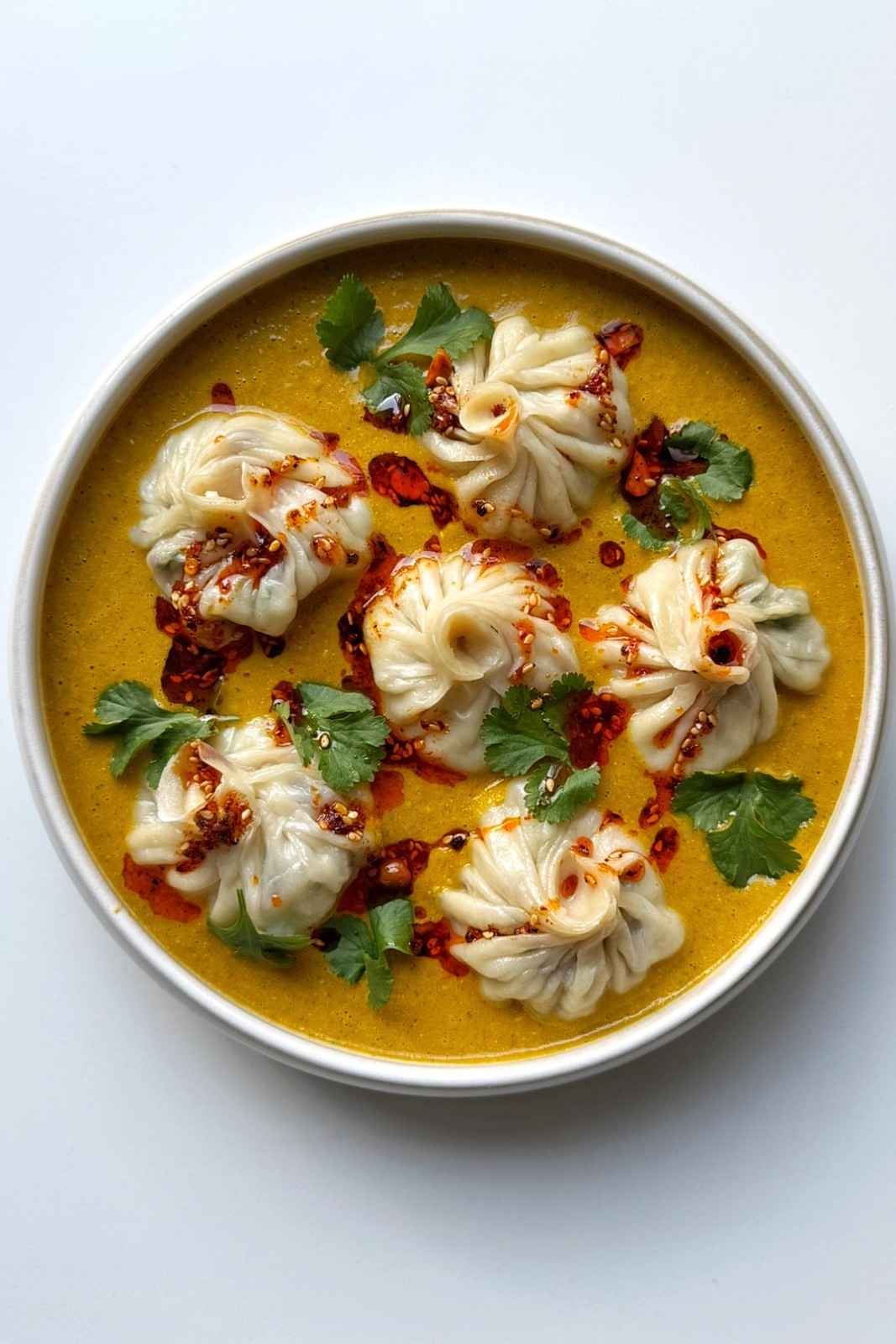 Overhead bowl of homemade jhol momo, Nepalese soup dumplings, sitting in a golden spiced tomato jhol broth, drizzled with chili oil and topped with fresh cilantro.