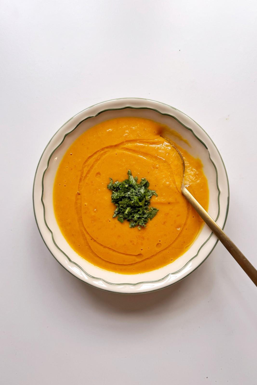 Overhead bowl of vegan carrot ginger soup, smooth and vibrantly orange, topped with fresh chopped cilantro and a gold spoon.