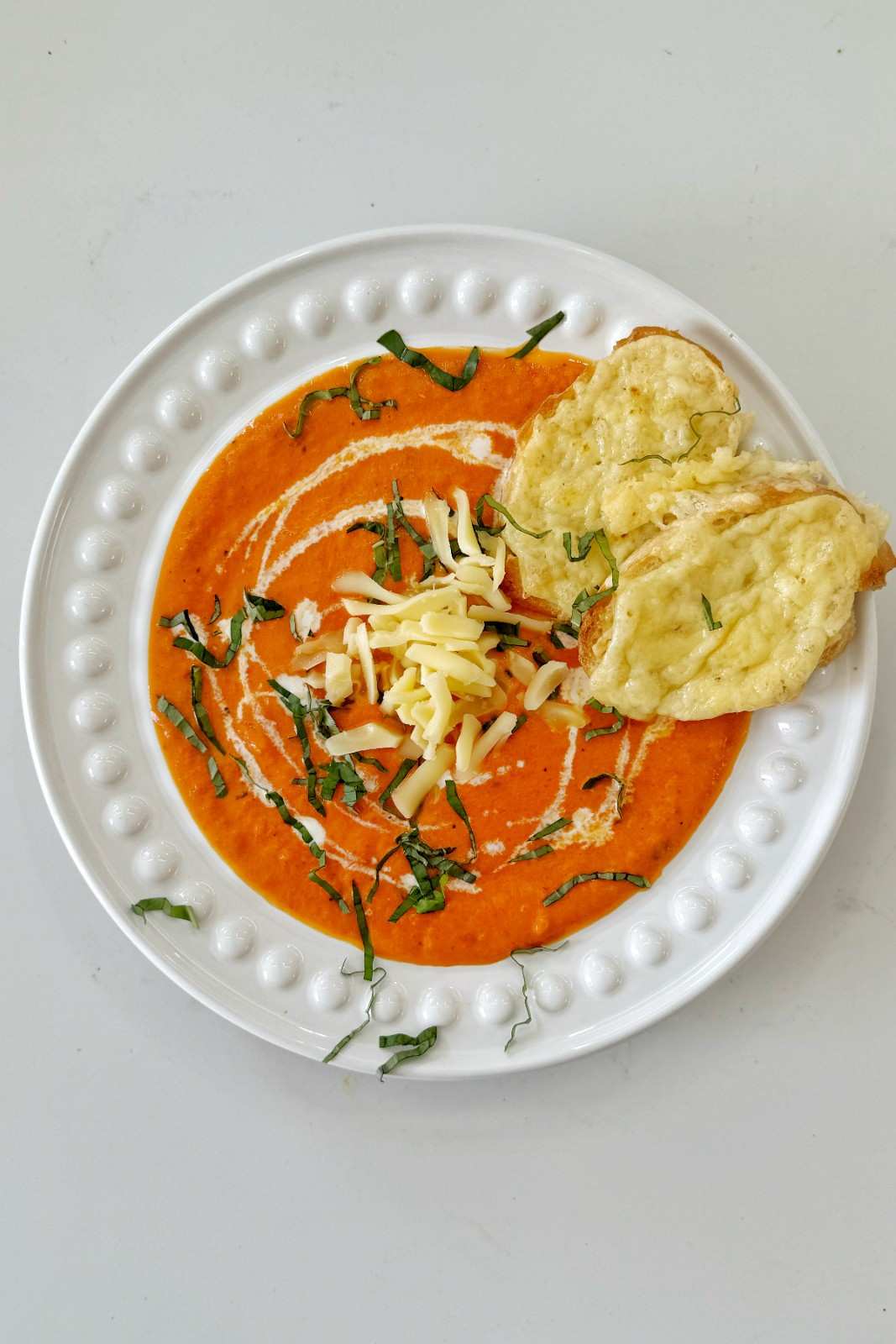 Overhead bowl of roasted red pepper and gouda soup, smooth and vibrant orange, topped with shredded gouda, fresh basil, a cream swirl, and a grilled cheese toastie on the side.