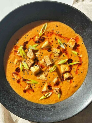 Overhead bowl of creamy tofu tomato soup topped with crispy tofu cubes, sliced scallions, chili oil, and black sesame seeds.