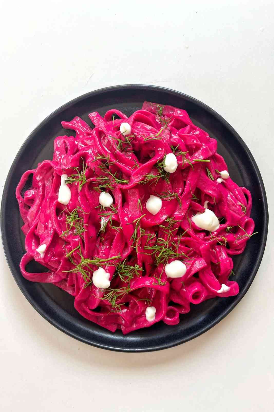 A top-down shot of long, wide tagliatelle pasta coated in a vibrant, bright pink beetroot and feta sauce, served on a black plate and garnished with small dollops of white cheese and fresh dill sprigs.