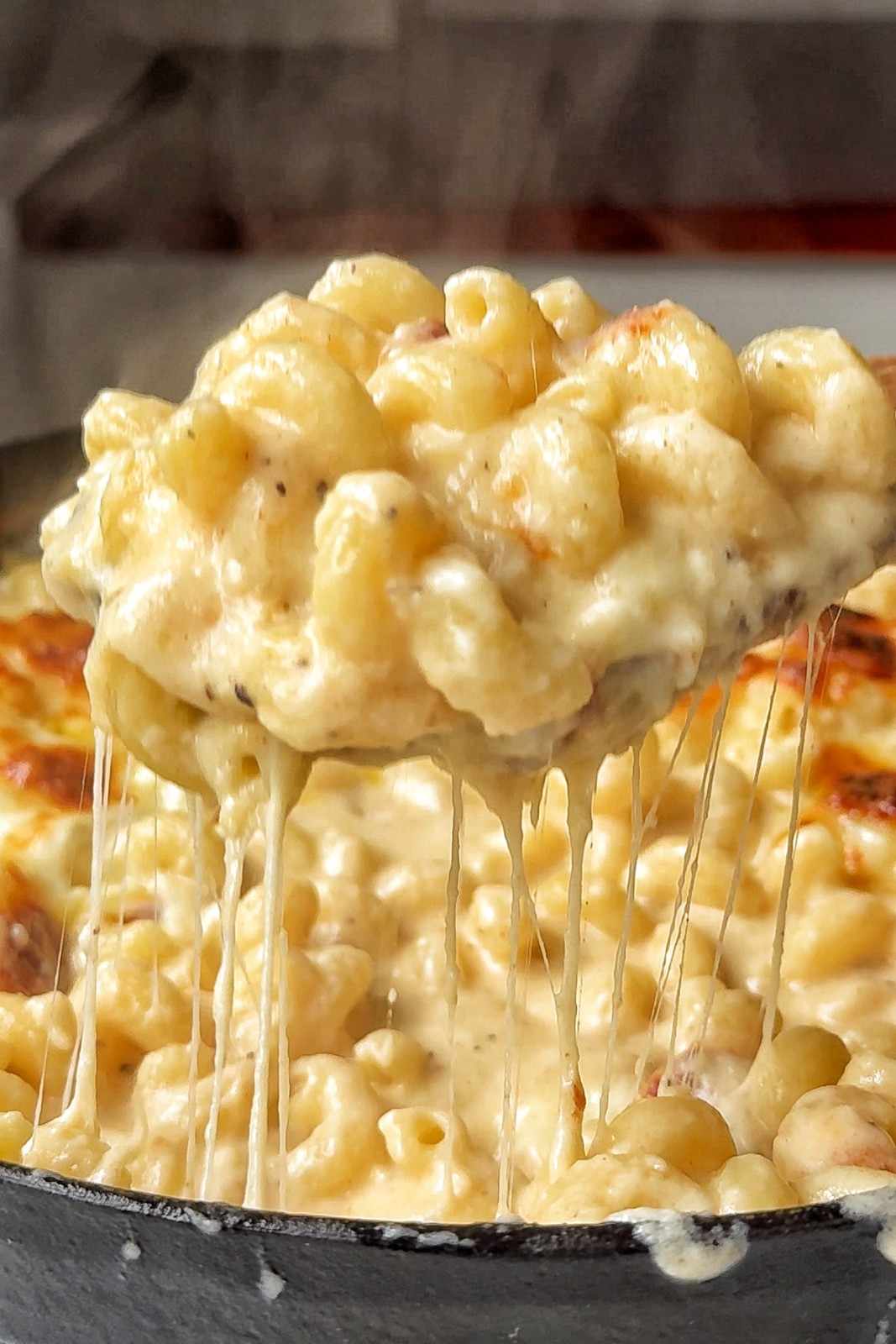 A close-up shot of a large spoonful of baked macaroni and cheese being lifted from a cast-iron skillet, creating long, dramatic strings of melted cheese stretching back into the creamy pasta dish below.