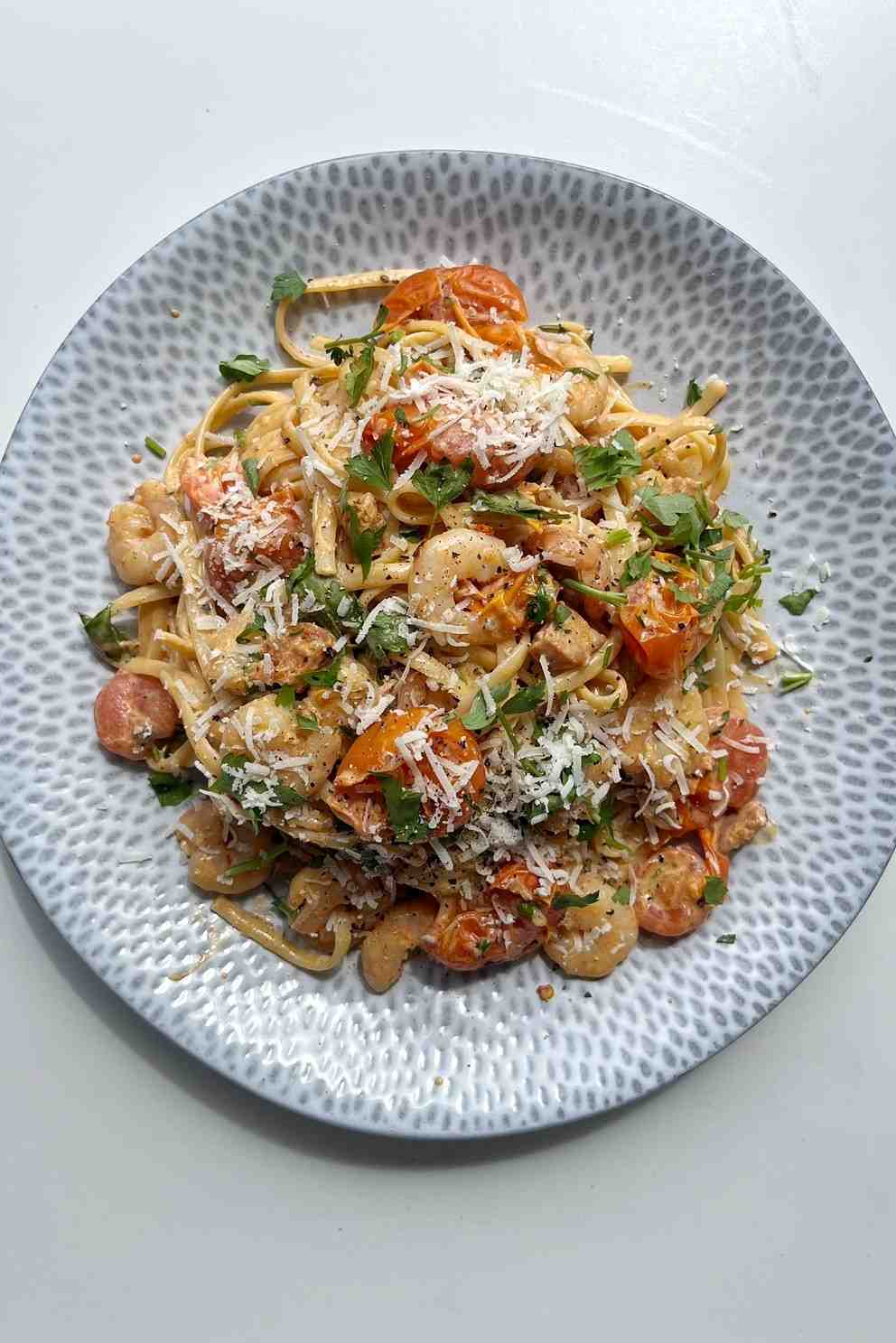 A top-down shot of a mound of linguine pasta tossed with juicy prawns, sliced chorizo, and burst cherry tomatoes in a creamy sauce, served on a grey patterned plate and topped with grated cheese and fresh parsley.