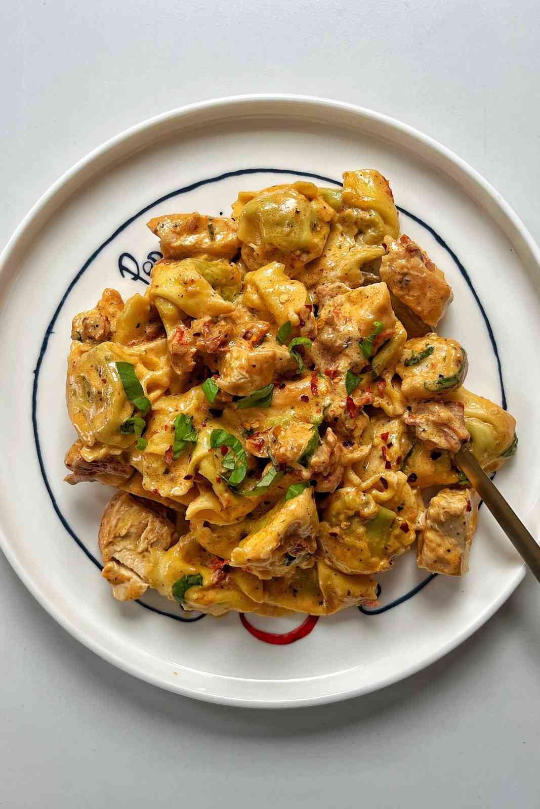 A top-down shot of plump tortellini and diced chicken coated in a rich, creamy sun-dried tomato sauce, served on a white ceramic plate with a thin blue rim and garnished with fresh torn basil and chili flakes.