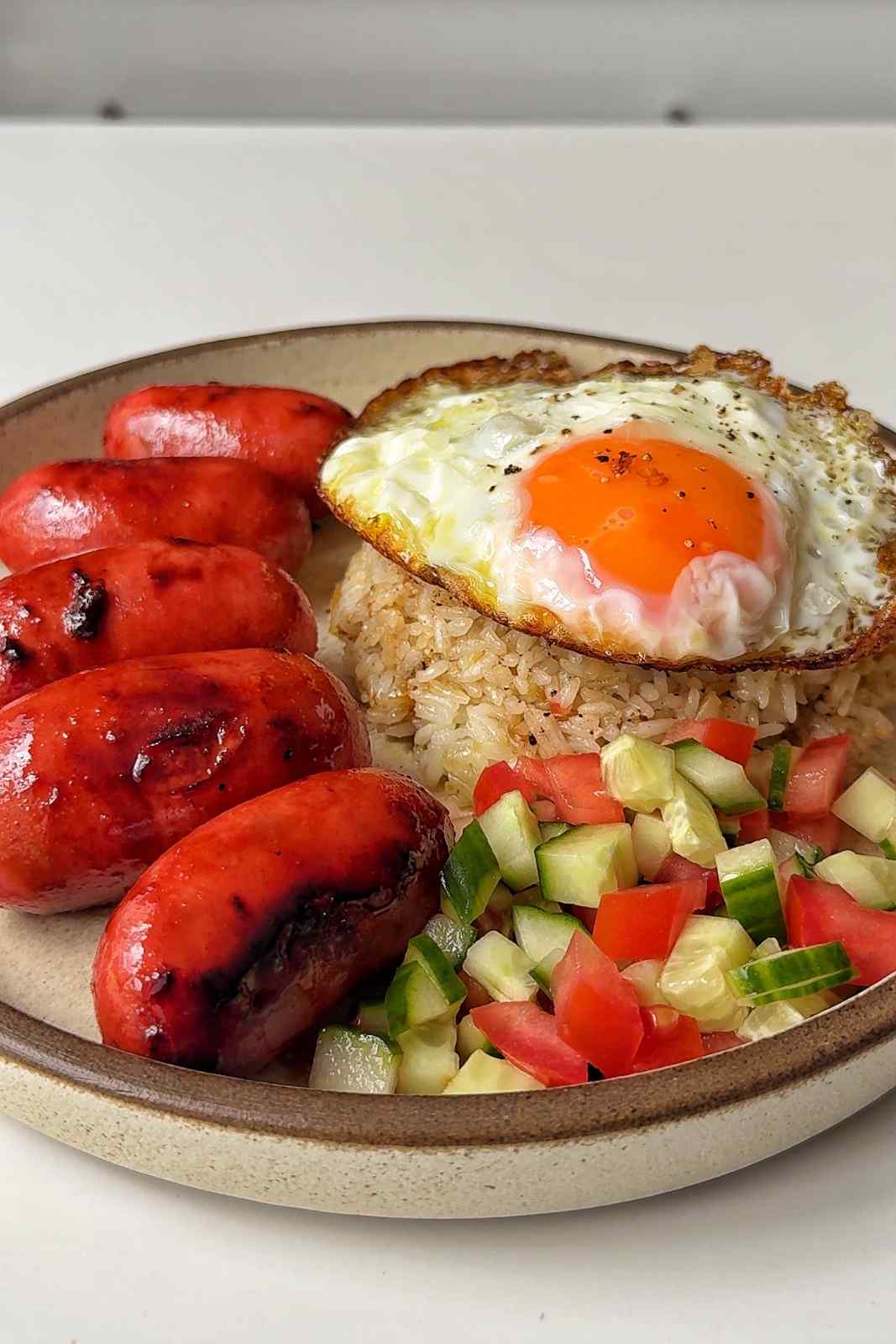 Longsilog on a plate with fried egg, garlic rice, fresh cucumber and tomato, and sausage.