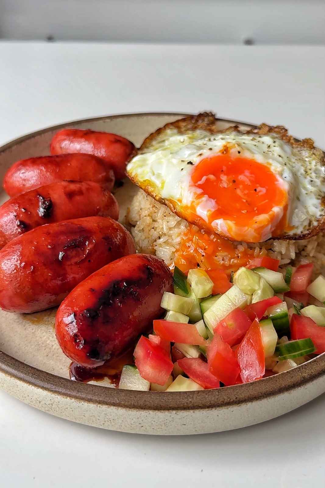 a plate of sausages, garlic fried rice, fried eggs, tomatoes, and cucumbers.