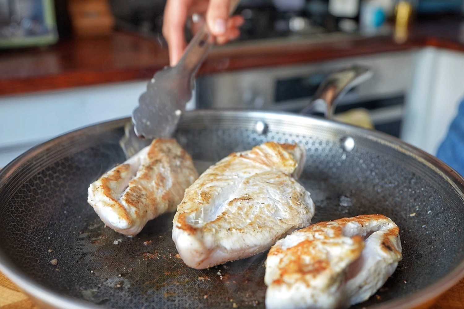 Three chicken breasts pan-frying in a stainless steel skillet, turned with tongs.