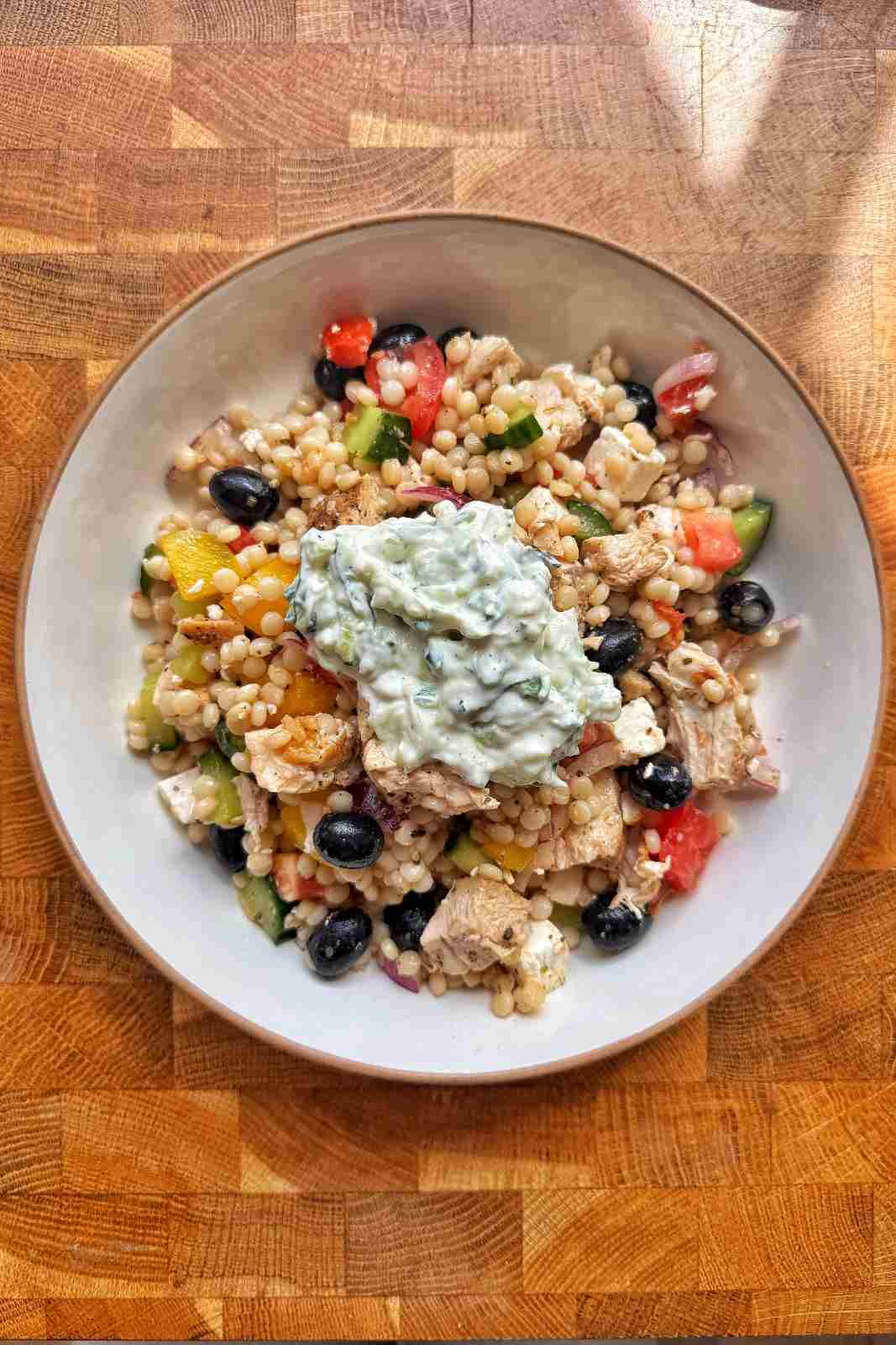 Overhead view of Greek pearl couscous salad in a bowl with chicken, feta, black olives and a dollop of tzatziki on top.