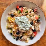 Overhead view of Greek pearl couscous salad in a bowl with chicken, feta, black olives and a dollop of tzatziki on top.