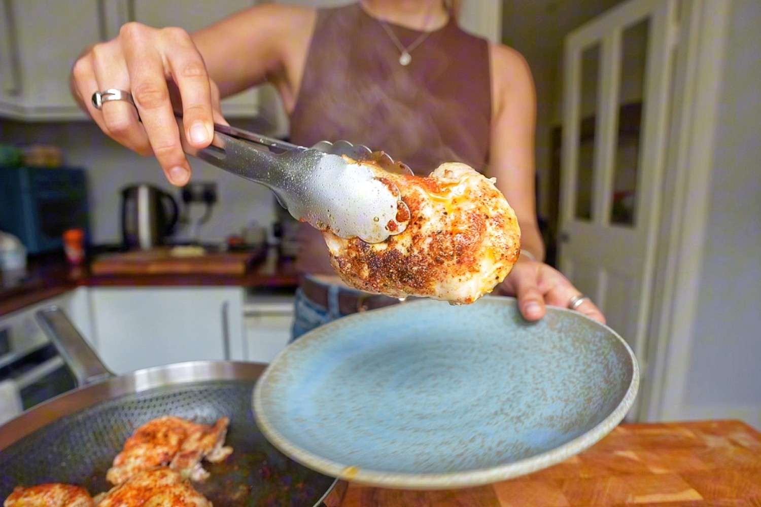 Emily lifting a seared chicken thigh out of a hot skillet with tongs onto a blue plate.