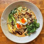 Overhead view of spicy peanut sausage ramen in a speckled bowl with udon, jammy egg, bok choy, cilantro and chili oil.