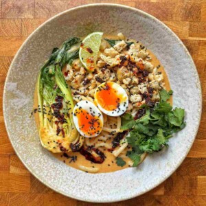 Overhead view of spicy peanut sausage ramen in a speckled bowl with udon, jammy egg, bok choy, cilantro and chili oil.