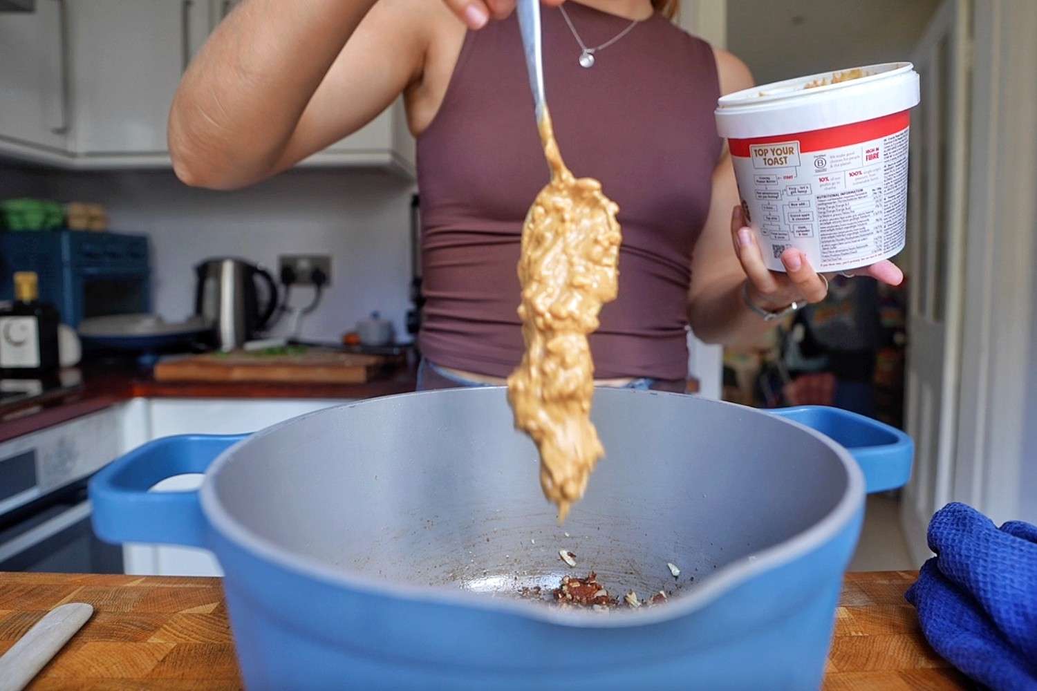 Spooning peanut butter into a blue pot with chopped garlic and Thai red curry paste.