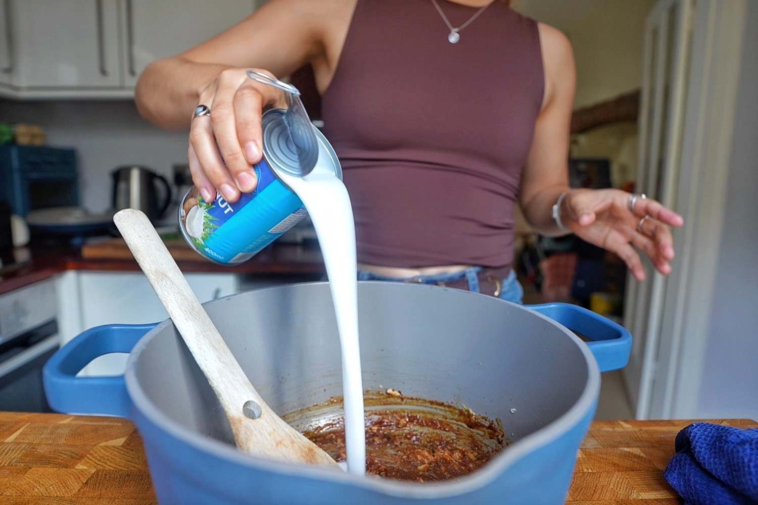 Pouring coconut milk from a can into a pot of Thai red curry paste and peanut butter.