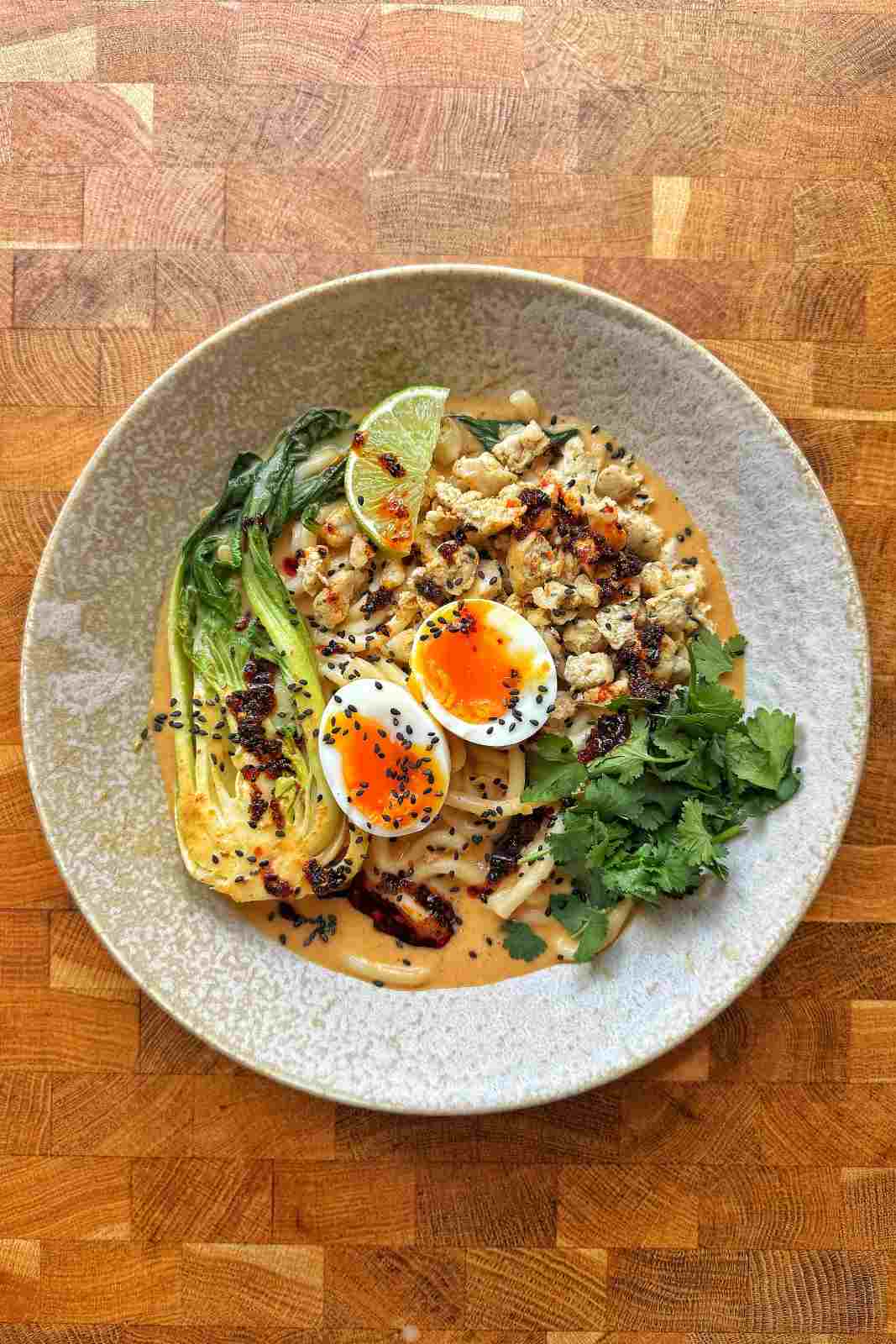 Overhead view of spicy peanut sausage ramen in a speckled bowl with udon, jammy egg, bok choy, cilantro and chili oil.