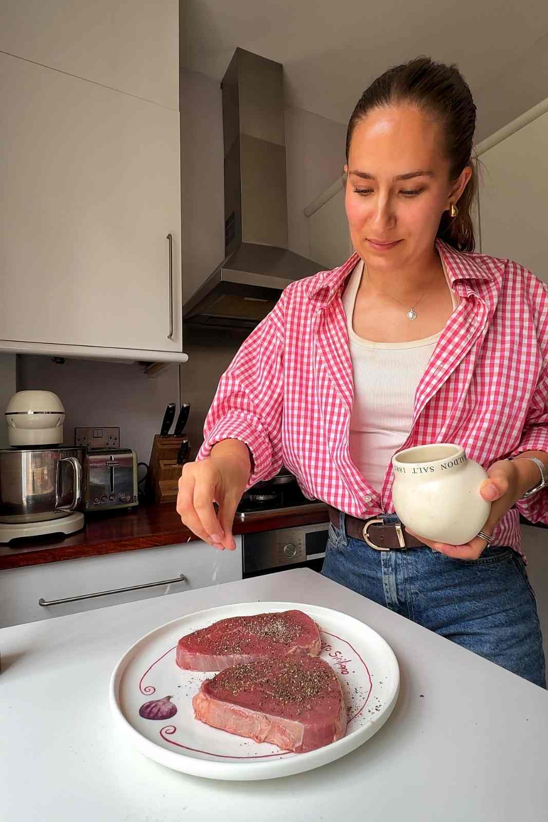 Emily seasoning two raw steaks with flaky salt and black pepper on a white plate.