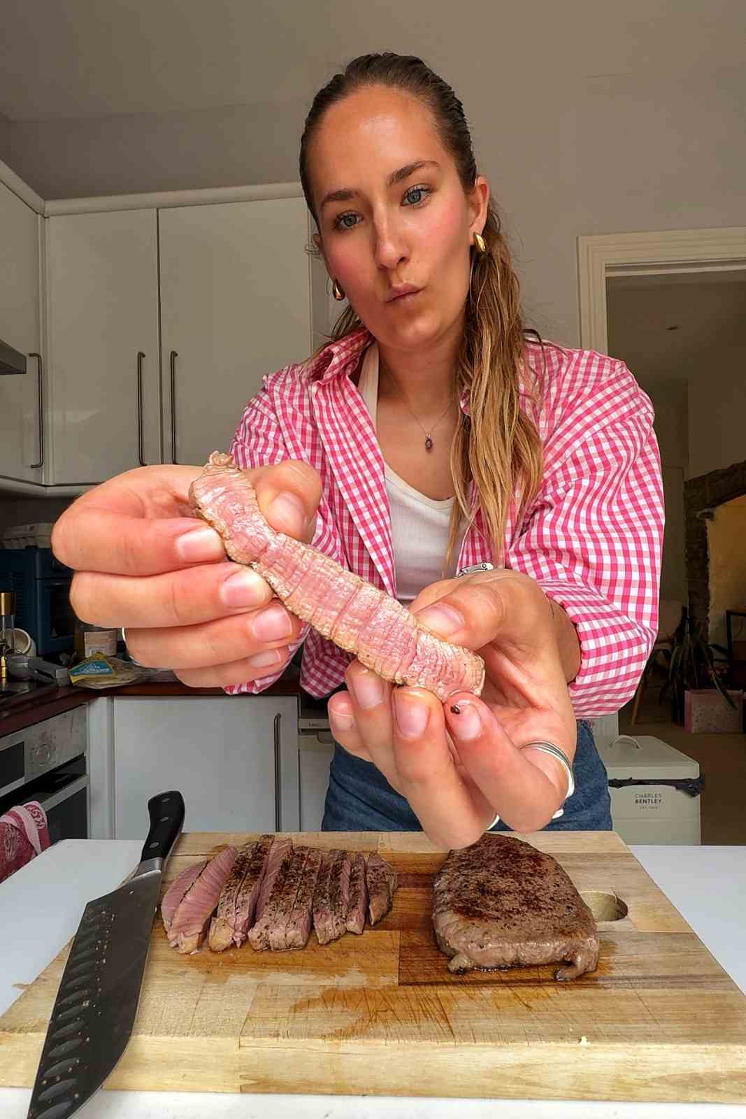 Emily holding a sliced steak showing a pink medium rare center next to a whole seared steak on a wooden board.