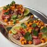 Two finished steak tacos on a silver platter topped with charred corn, pickled onion, cilantro and lime.