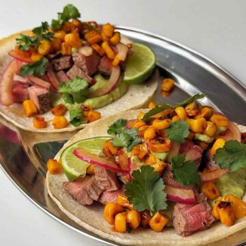 Two finished steak tacos on a silver platter topped with charred corn, pickled onion, cilantro and lime.