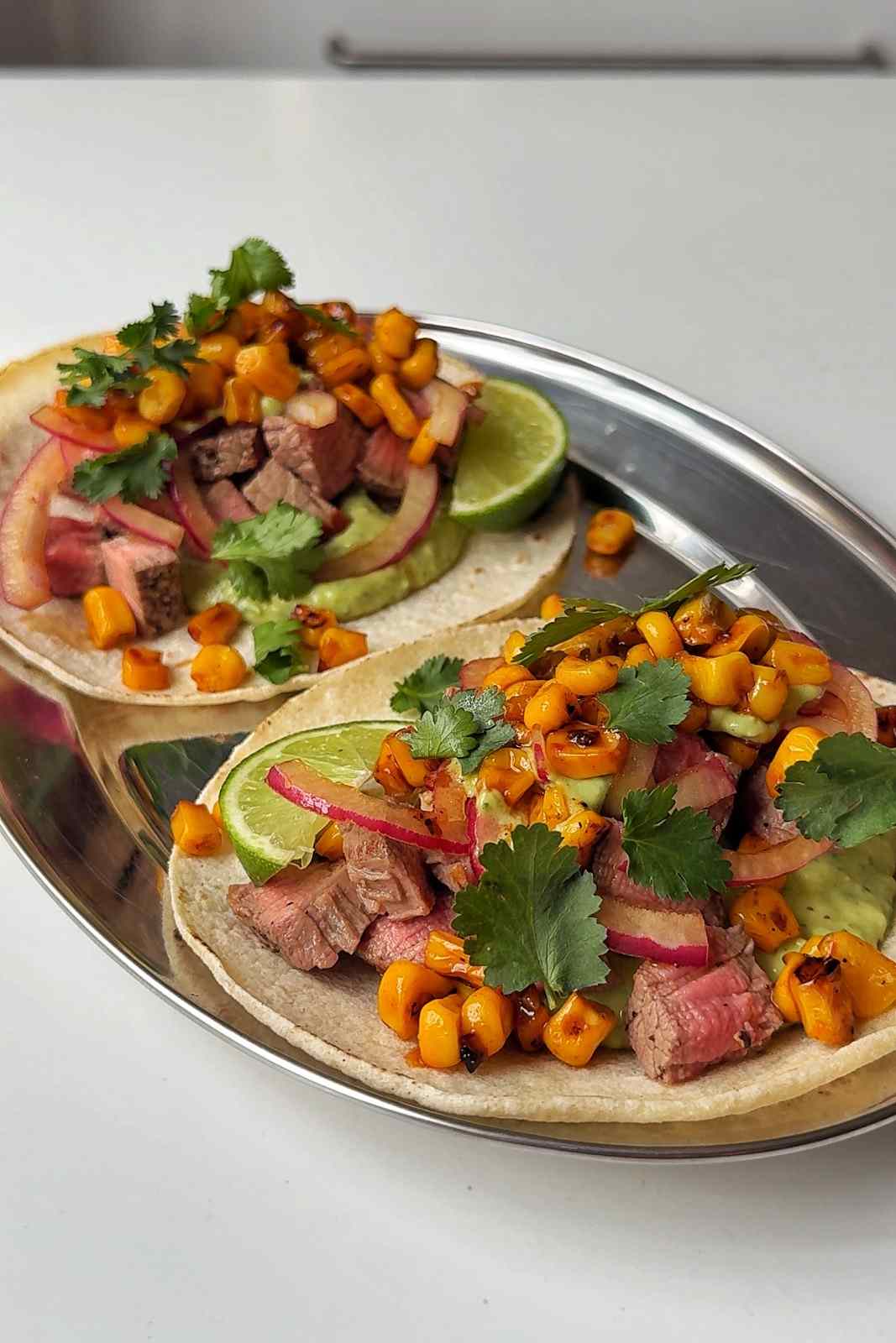 Two finished steak tacos on a silver platter topped with charred corn, pickled onion, cilantro and lime.