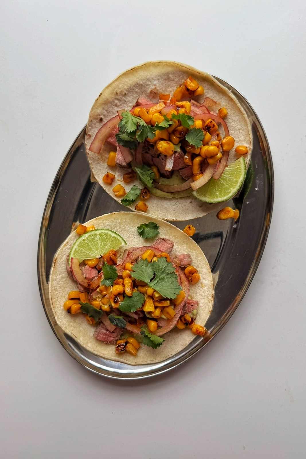 Overhead view of two steak tacos on a silver platter with lime wedges and charred corn.
