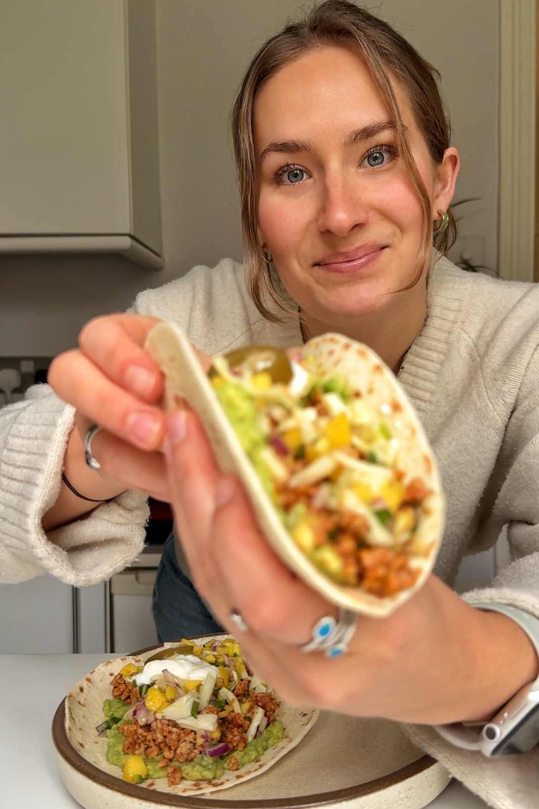 Emily holding a ground pork taco topped with mango salsa, sour cream and smashed avocado in her kitchen.