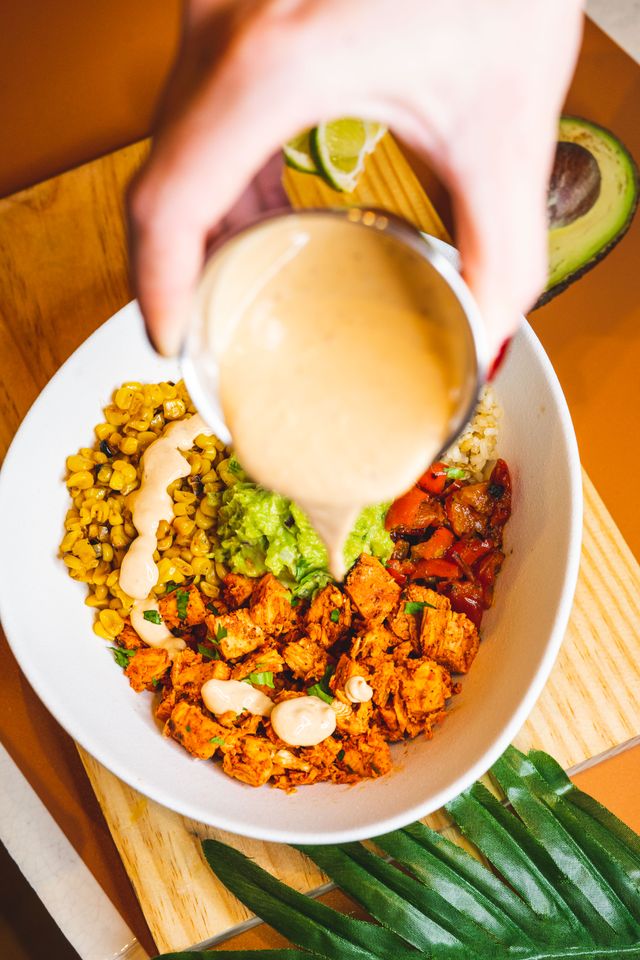 Creamy chipotle crema being poured over a bowl of spiced chicken, charred corn, guacamole and roasted red peppers.