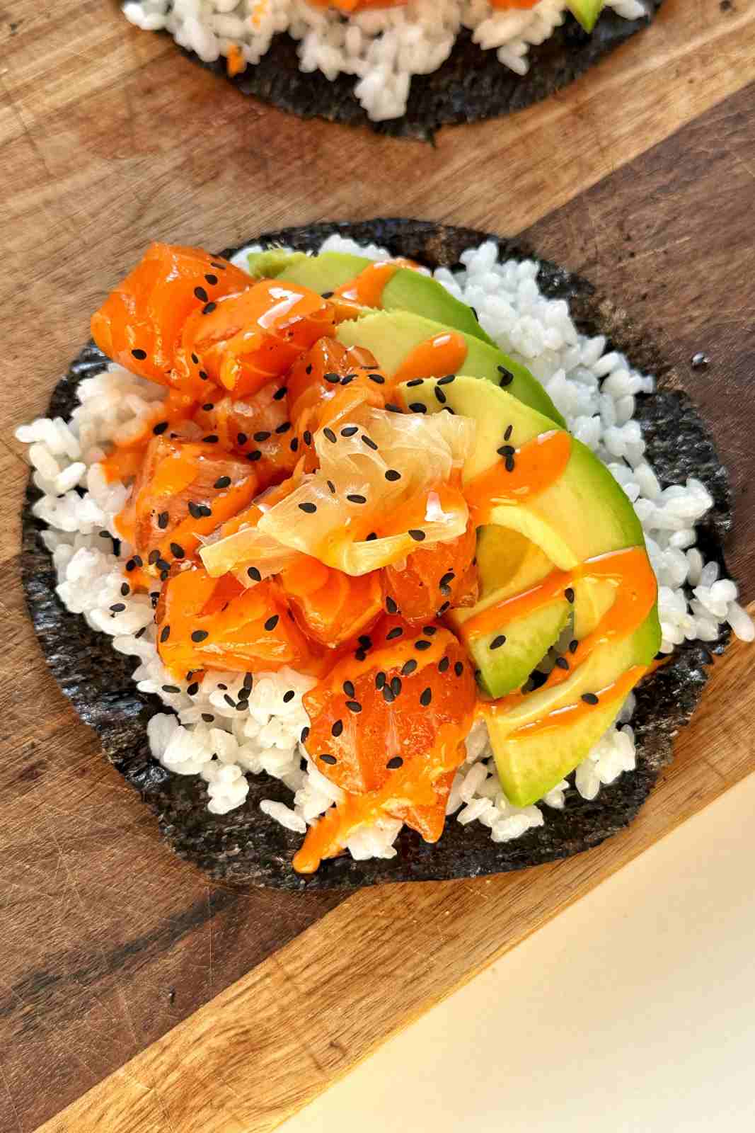 Spicy sriracha mayo drizzled over a sushi taco with raw salmon, avocado, pickled ginger and black sesame seeds on a round nori sheet.