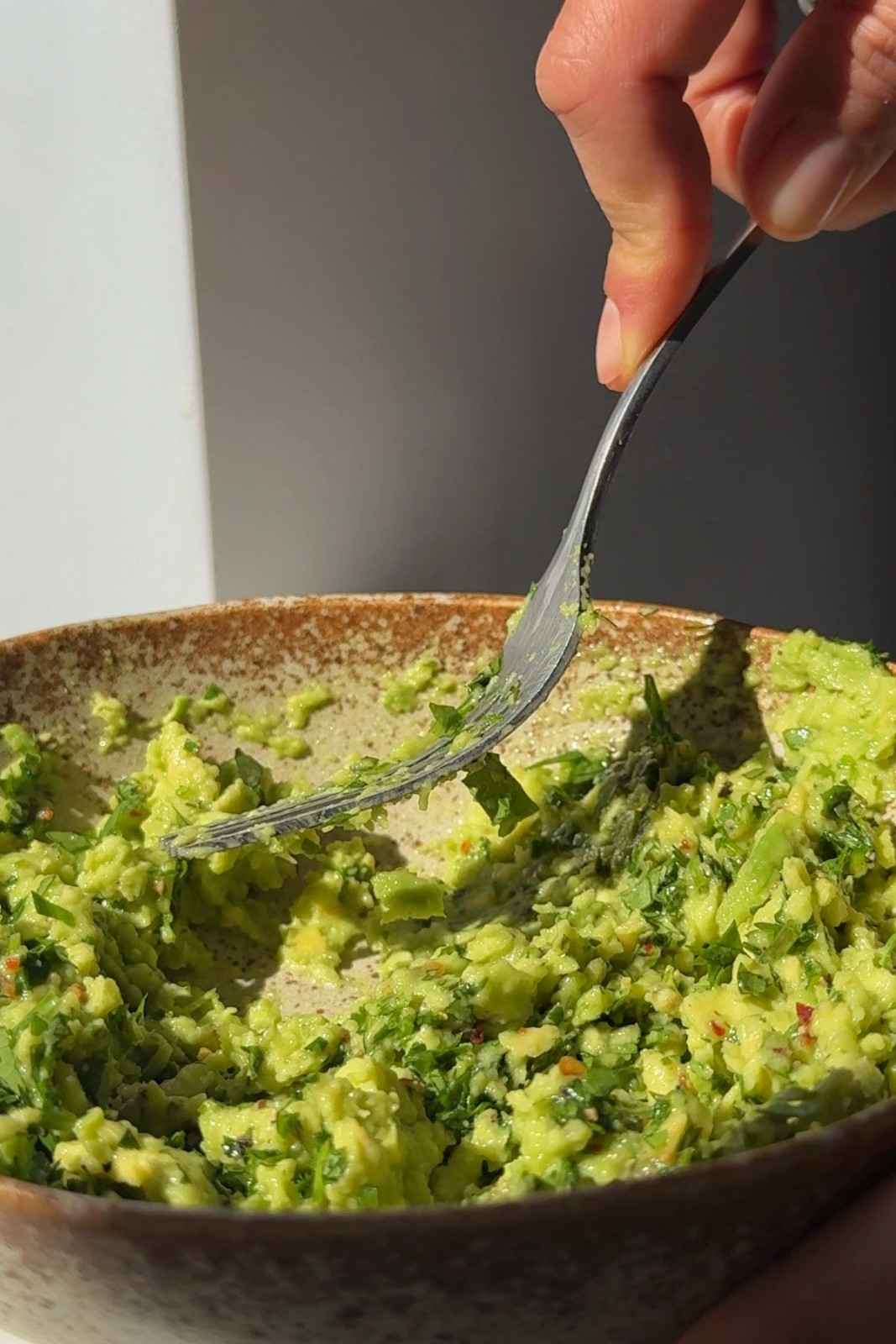 Guacamole being mashed with a fork in a speckled ceramic bowl with chopped cilantro and chili flakes