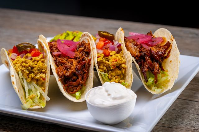 A bowl of sour cream served alongside four soft tacos with shredded meat, lettuce, pickled red onions and jalapeños.
