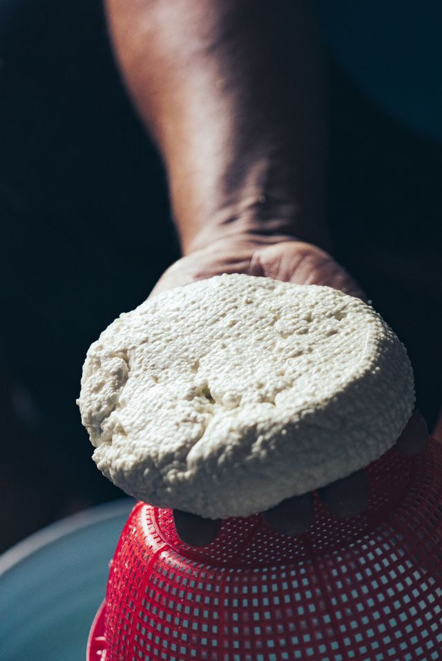 A hand holding a freshly made wheel of white queso fresco above a red draining basket.