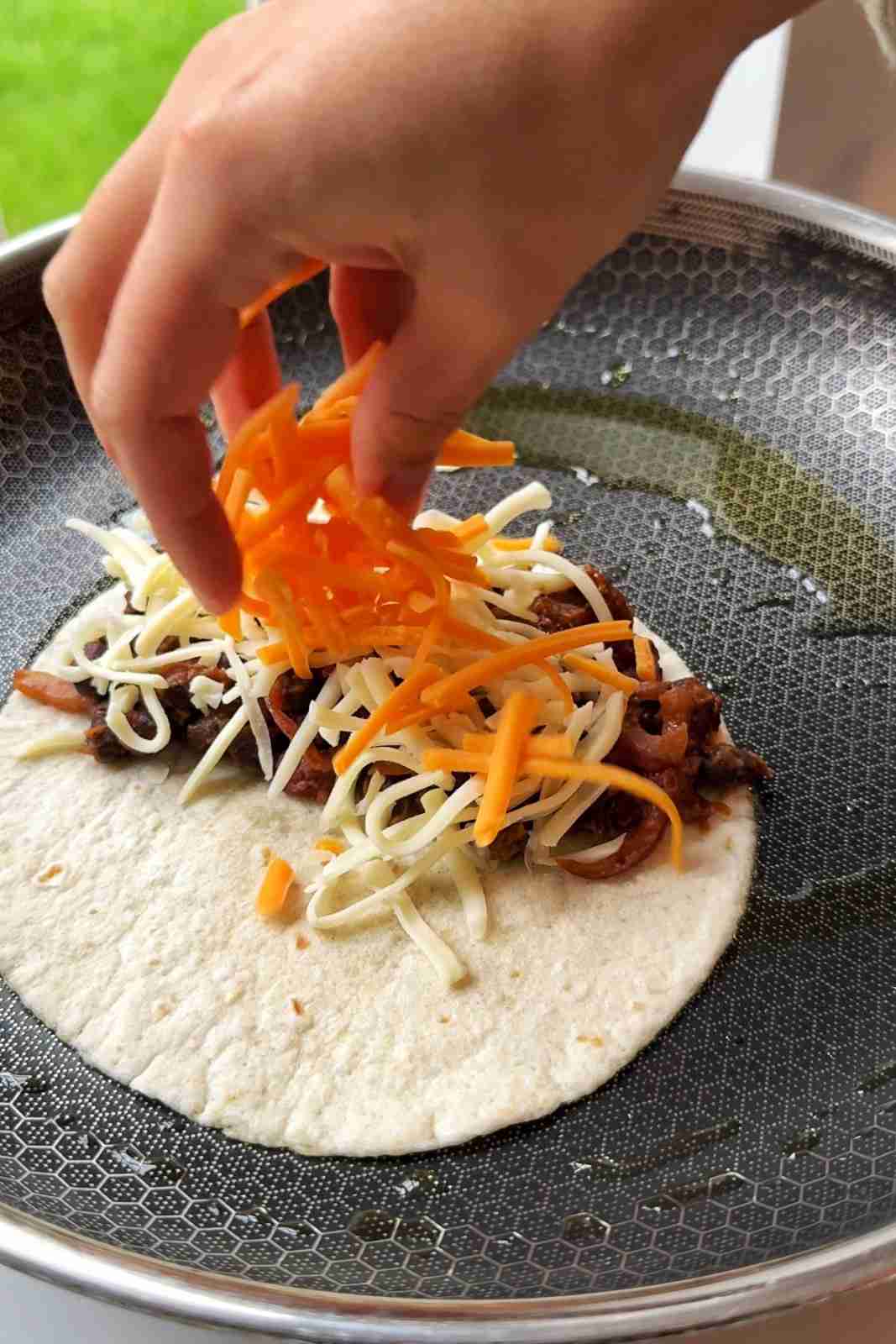 Scattering shredded cheddar and mozzarella over black bean filling on a soft tortilla in a frying pan.
