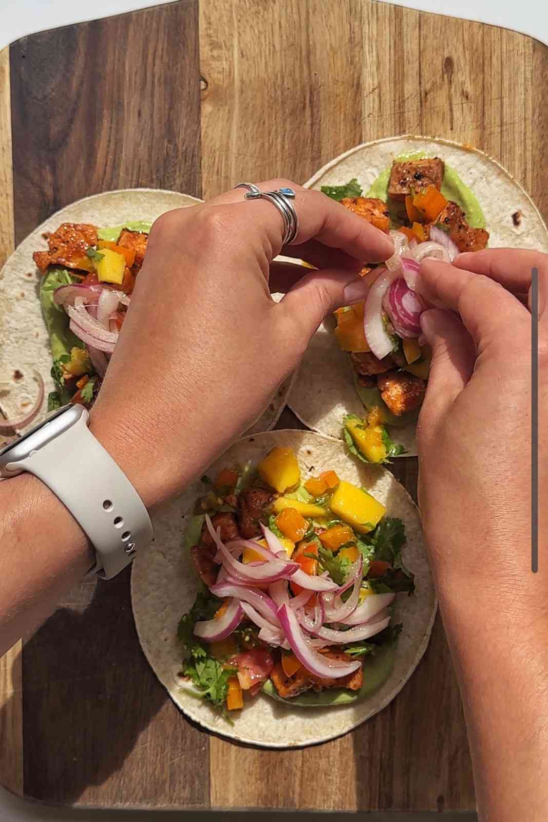 Placing pickled red onions onto tacos topped with mango salsa, avocado sauce and cilantro on a wooden board.