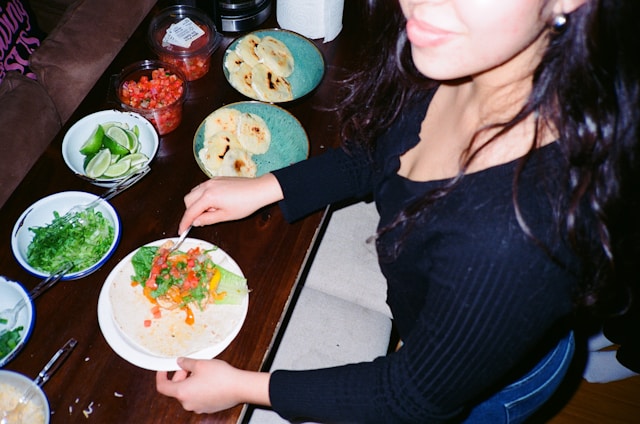 A bowl of fresh lime wedges on a taco bar spread with pico de gallo, shredded lettuce and warm tortillas as someone builds a taco.