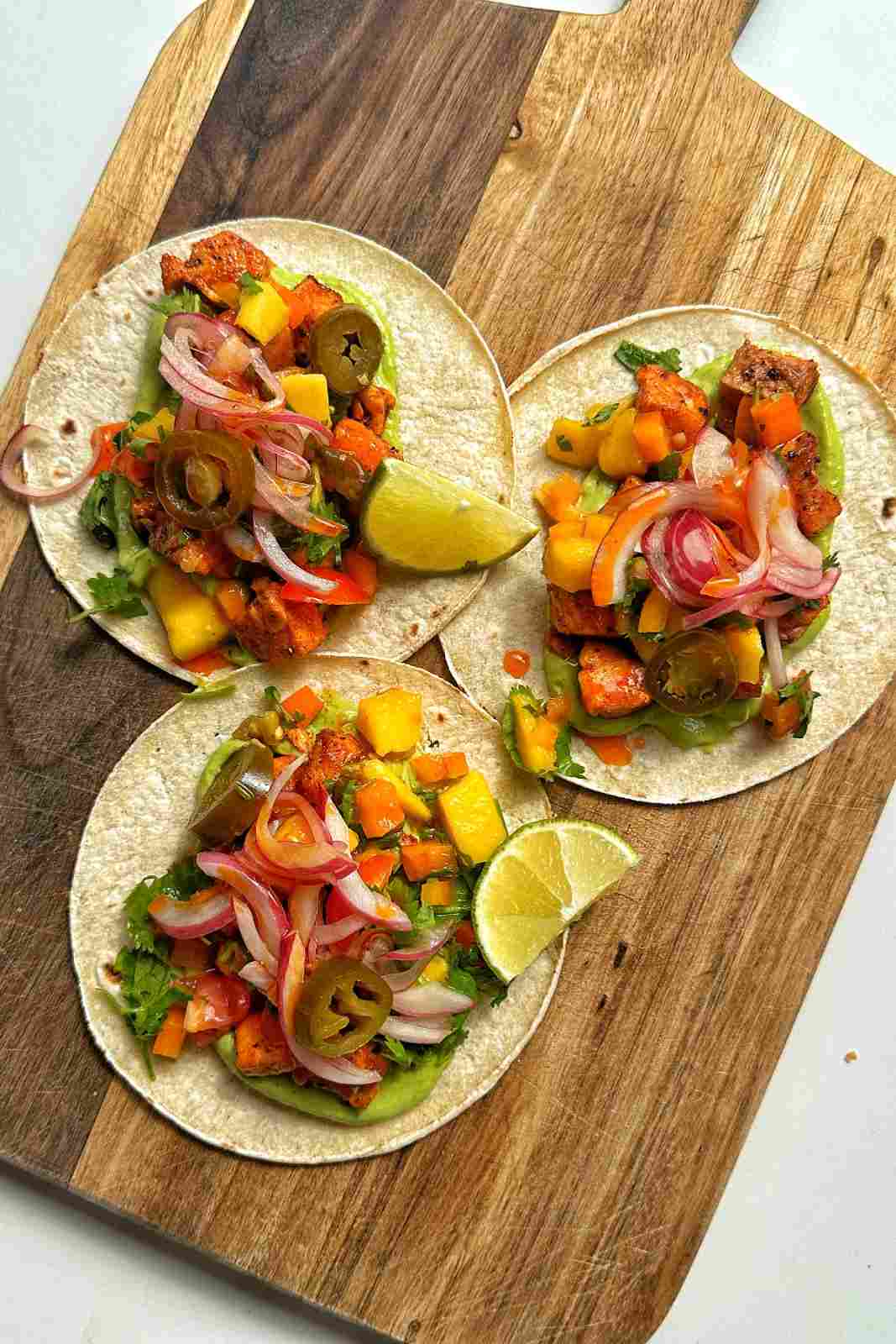 Three tacos topped with creamy green salsa and a drizzle of red salsa, with mango, pickled red onions, jalapeños and cilantro on a wooden board