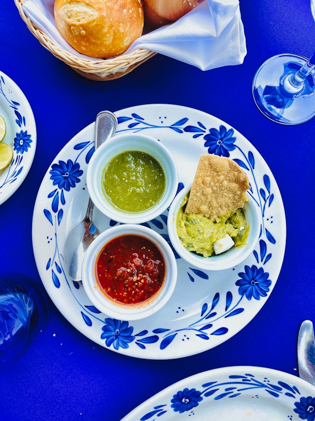 A ramekin of bright green salsa verde on a blue and white Talavera plate beside red salsa and guacamole with a tortilla chip.