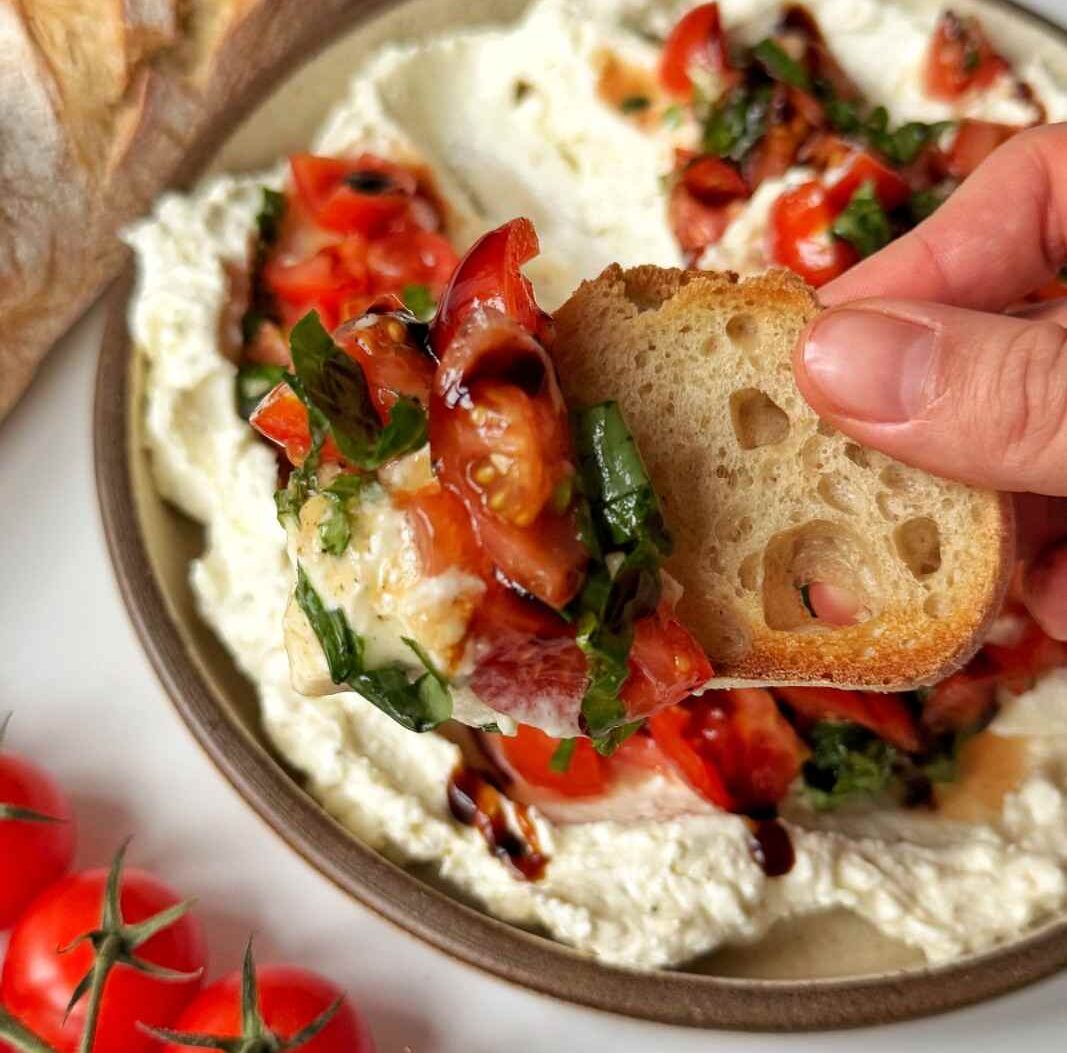 Emily scooping a whipped cream cheese and feta bruschetta topping with baguette, with the dip and tomatoes lathered onto the baguette in a thick layer. 